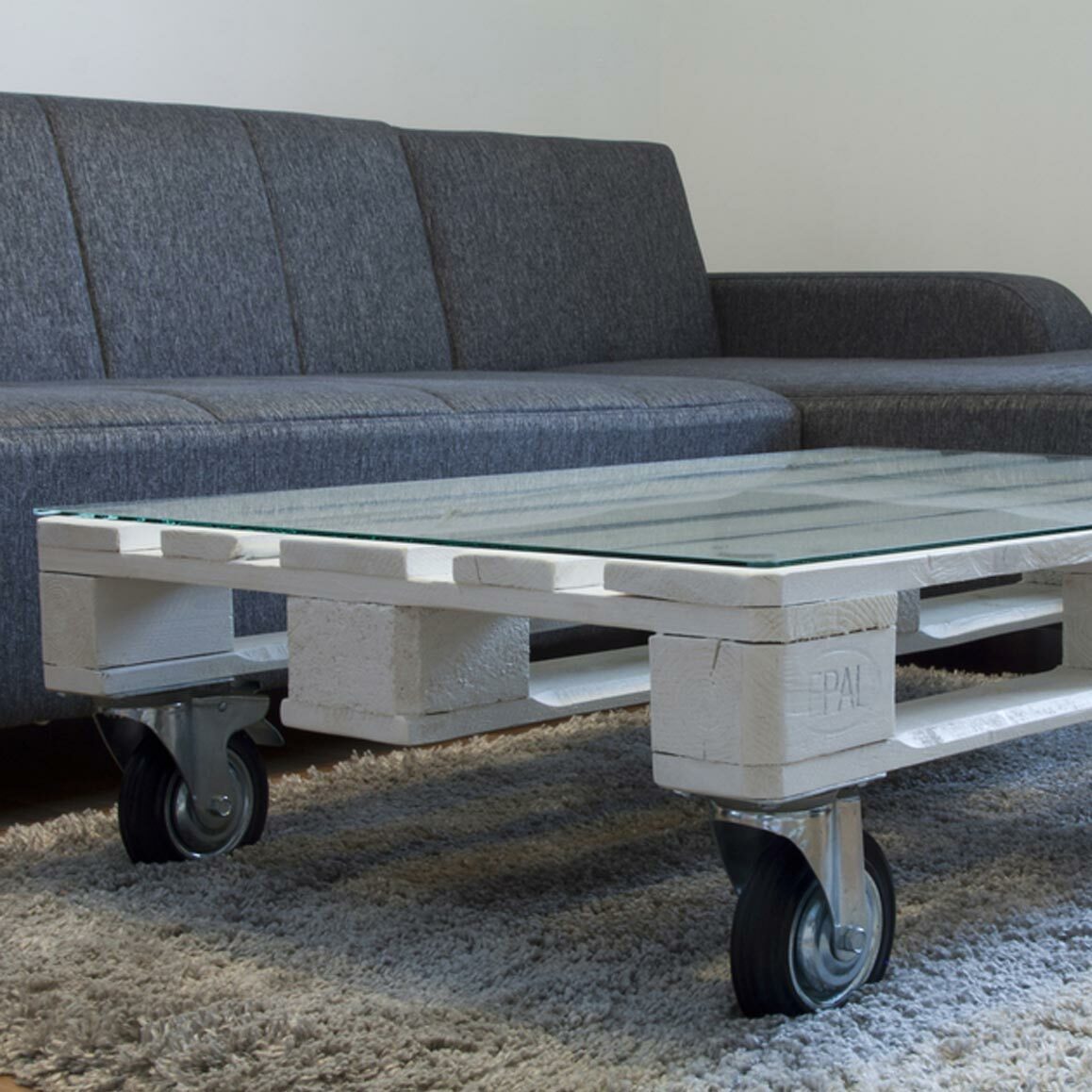 A modern living room featuring a gray couch and a unique coffee table made from a white wooden pallet with a glass top. The table is set on caster wheels and rests on a textured beige rug.