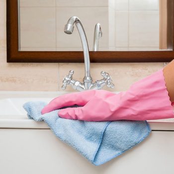 In the bathroom, hand in rubber protective glove cleaning with microfiber cloth washing and polishing ceramic sink and water tap