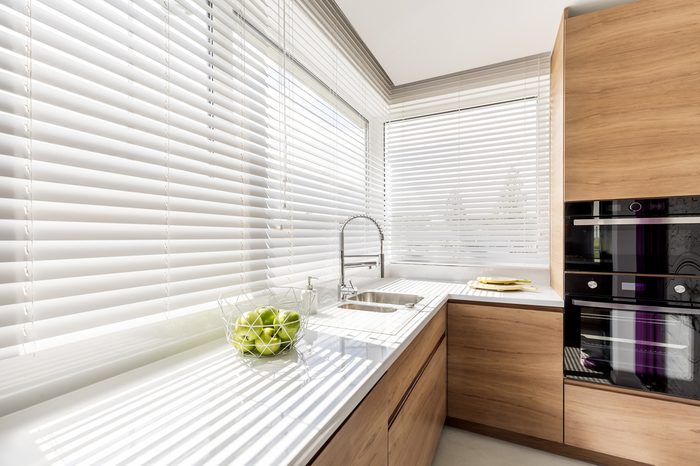 Modern bright kitchen interior with white horizontal window blinds, wooden cabinets with white countertop and household appliances