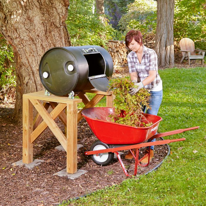 woman adding leaves from a whellbarrow to a homemade Drum Composter in a backyard under a large tree