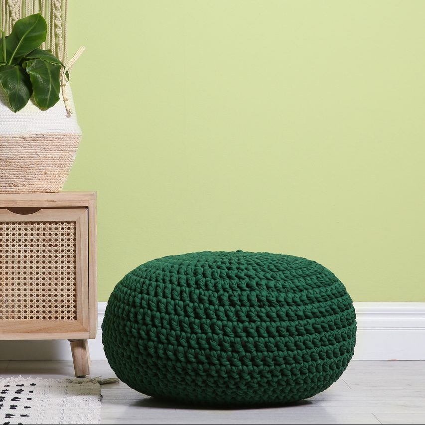 Stylish room interior with comfortable knitted pouf, wooden furniture and plant near light green wall, space for text