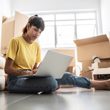 Young woman using laptop on the floor in search of a new apartment with moving boxes in the background