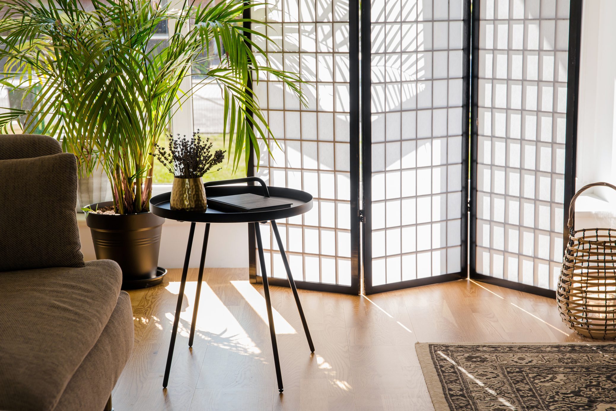 Home living room with wood and paper divider screen blocking sun from window, black metal accent table and sofa, natural colors.