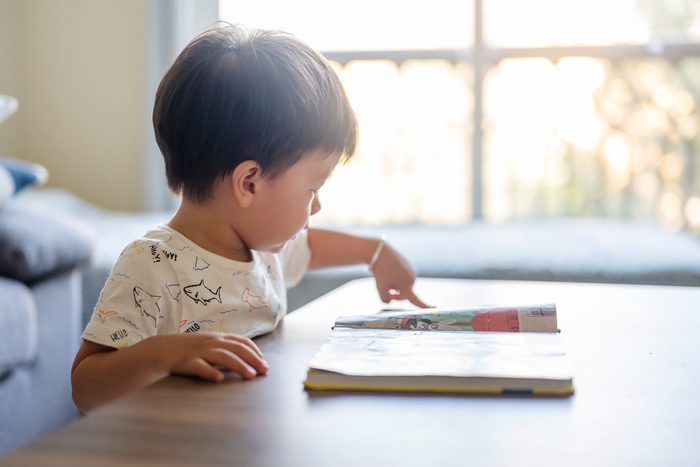 Baby boy looking at a book standing by a coffee table in the living room at home