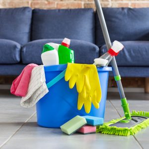 Bucket with bottles of detergent, cleaning tools and mopping stick in front of sofa in living room. Housework cleaning equipment.