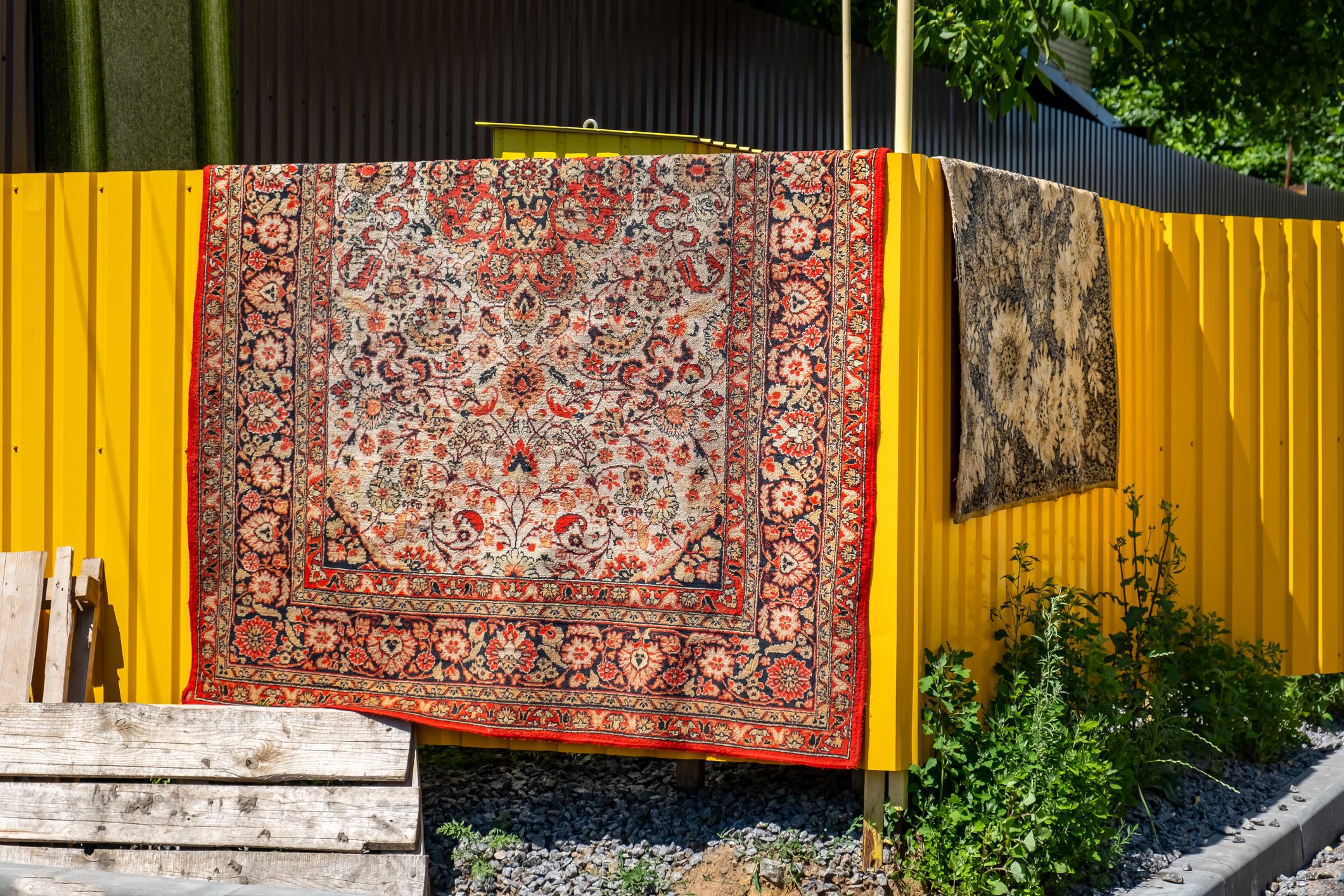 An Oriental red carpet with patterns dries on a yellow metal fence near the house. Carpet cleaning and drying.