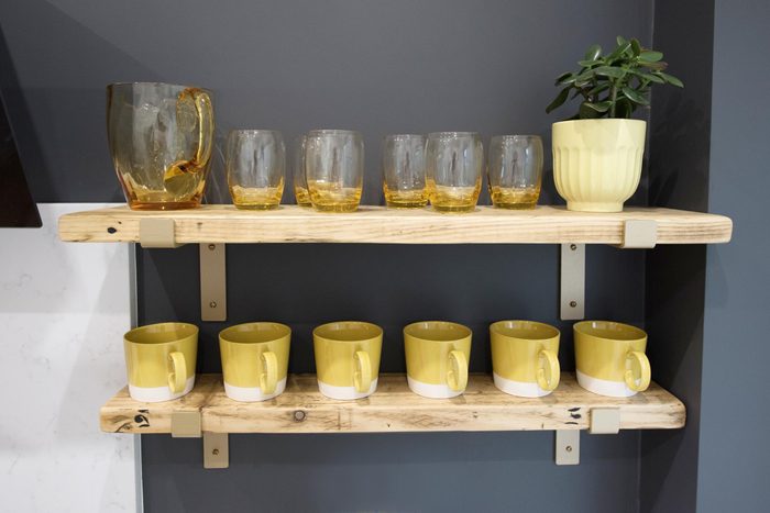 Yellow cups, amber coloured glass drinking glasses and jug and potted plant are displayed on wooden shelves made from reclaimed scaffold boards supported by gold colored metal brackets