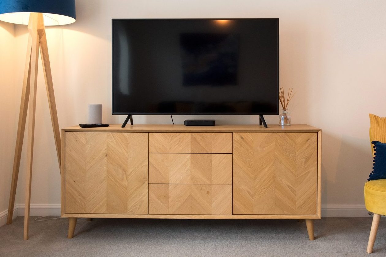 A general interior view of a lounge with yellow accent chair with navy blue cushion, sideboard with flat screen television set, floor standing tripod style lamp