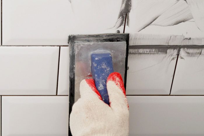 Workers' Hands Using A Rubber Spatula And Grouting With Paste Between White Ceramic Tiles to grout seams in a tile bathroom backsplash