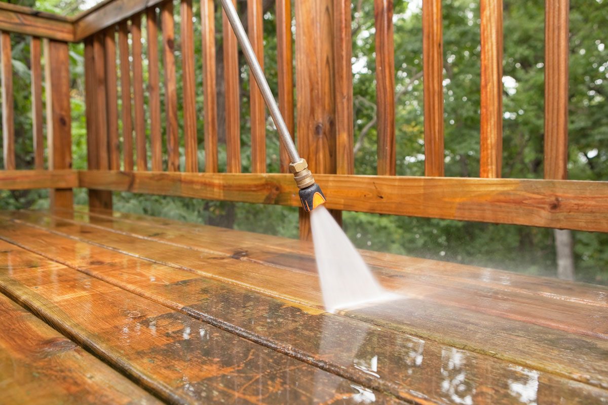 A pressure washer sprayer is cleaning a weathered treated wood deck.