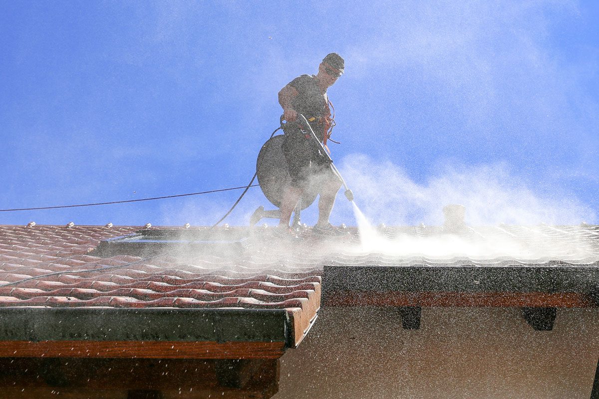 Worker secured with a safety rope cleaning the roof of moss and dirt with a high pressure washer with pressurized water