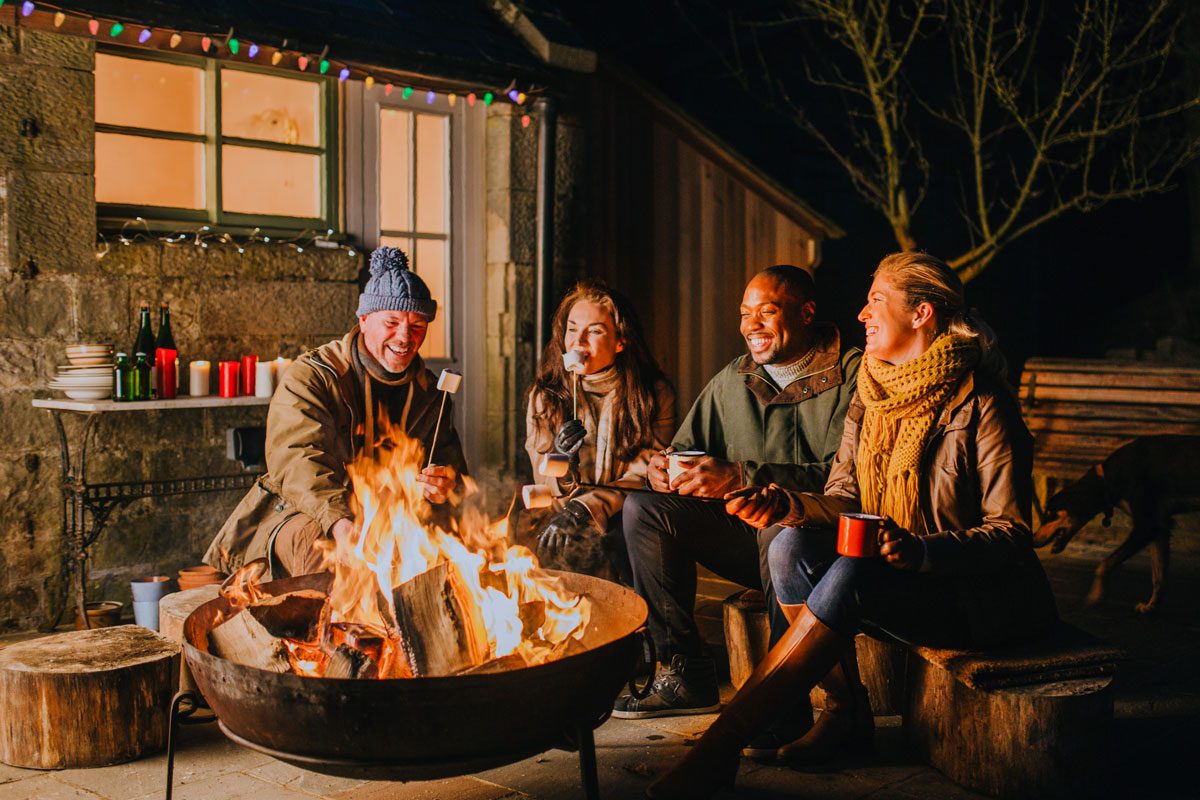 Group of mature friends in their 30s and 40s gathered around a fire pit in a garden.