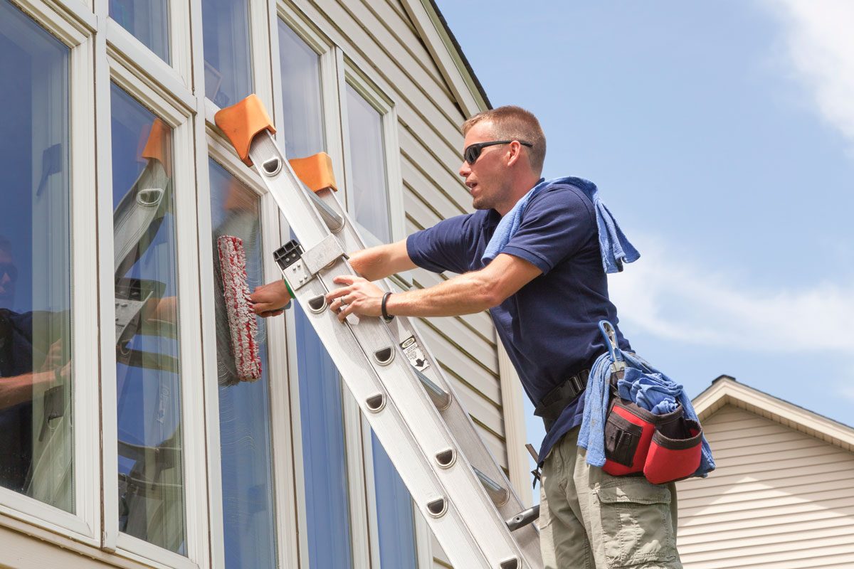 Young Business Man Window Washing Professional Washes Windows on a Residential Home with a ladder, cleaning sponges and proper safety equipment on a blue, sunny day