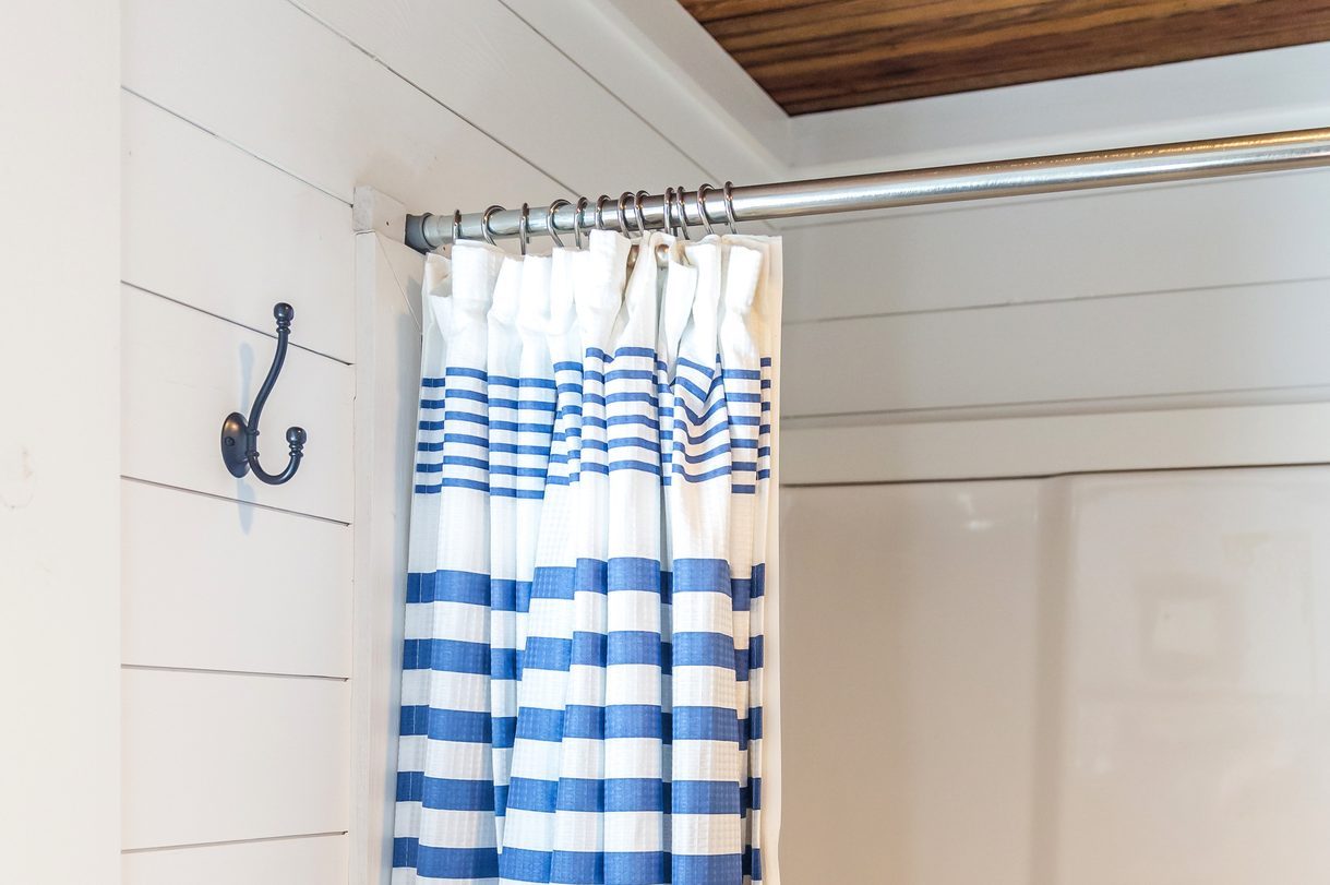 Small and compact bathroom shower stall in a small farmhouse decor inspired cabin with a blue striped shower curtain, hooks on the wall and a rustic wood plank ceiling