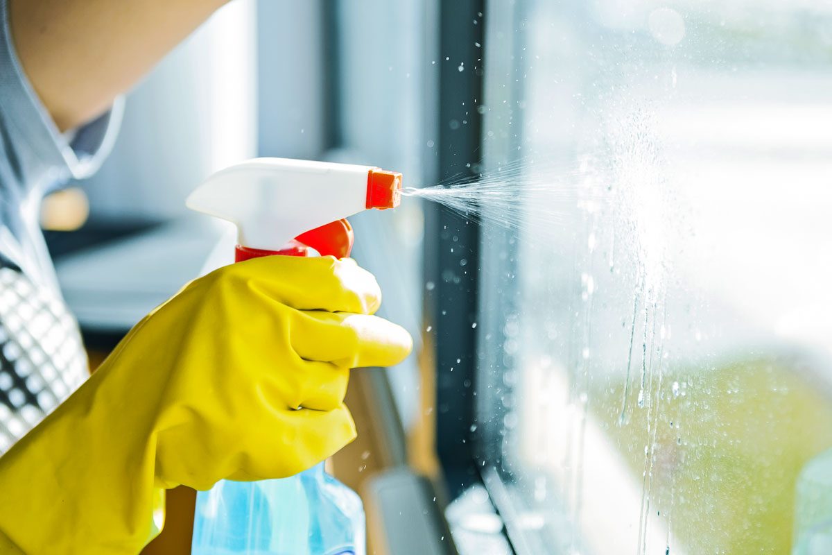 Young Woman Washing Windows Using Glass Cleaning Spray, Yellow Rubber Gloves, against a brightly lit window sill