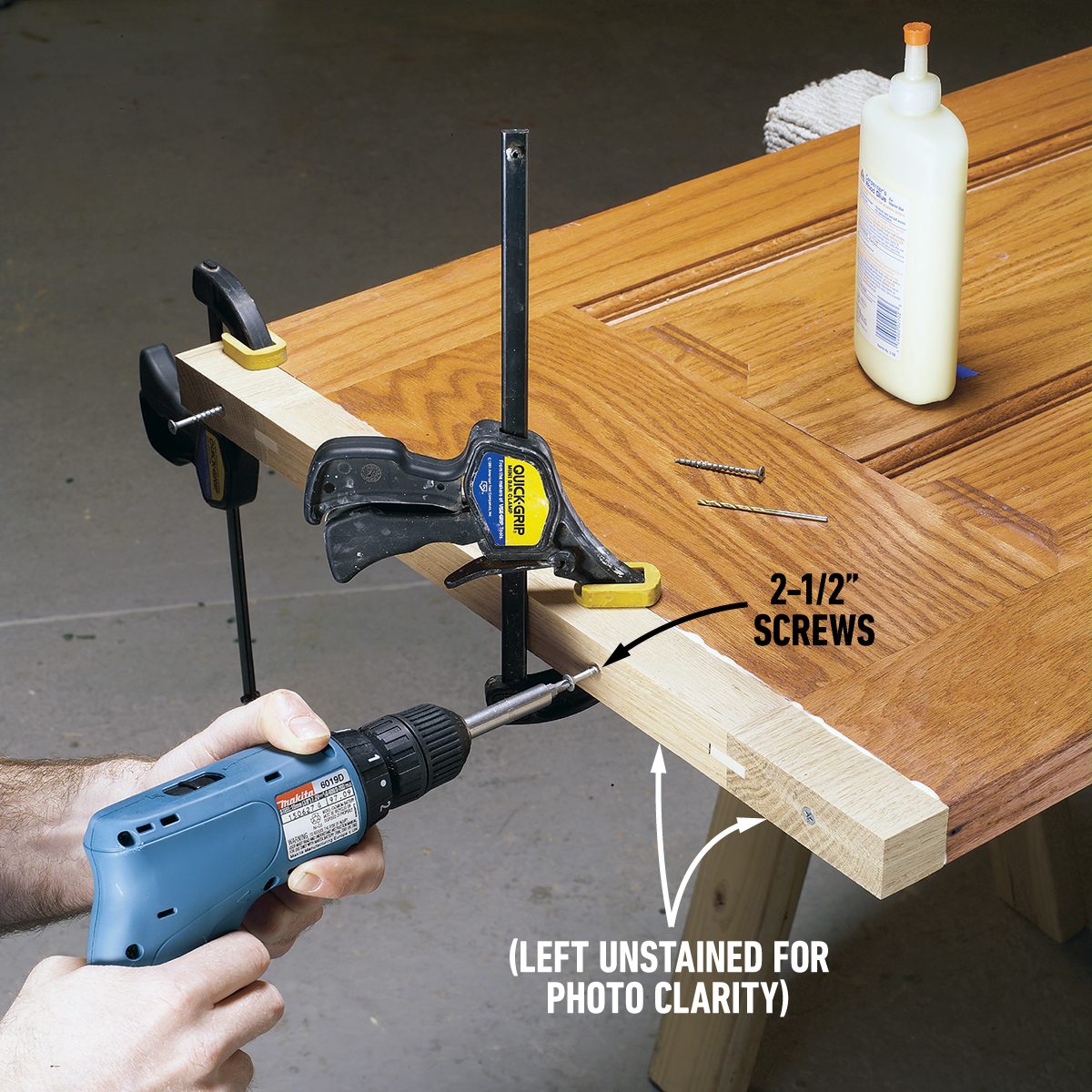 A person uses a blue power drill to drive a screw into a piece of wood, which is clamped to a wooden work surface. The wood is labeled "left unstained for photo clarity." Nearby are wood glue and two nails. The setup demonstrates a woodworking assembly.
