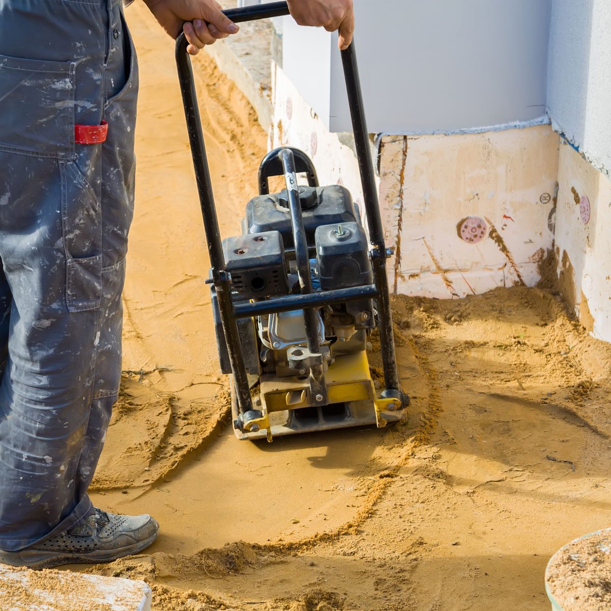 Worker Compresses Sand In Blind Area Around Building With Tamping Machine