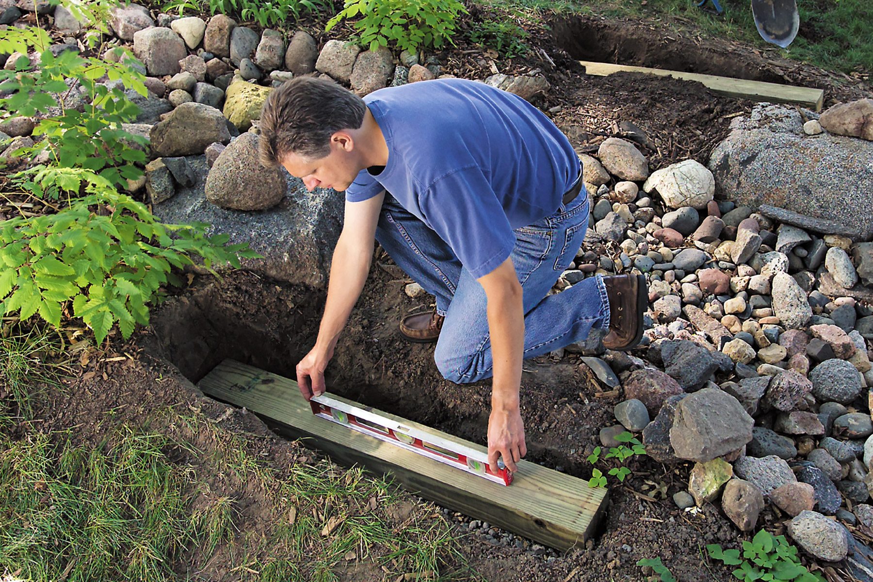 A person kneels next to a dug-out patch of soil, placing a long wooden beam within it. They are using a spirit level to ensure the beam is even. Surrounding the individual are various rocks, plants, and other landscaping elements.