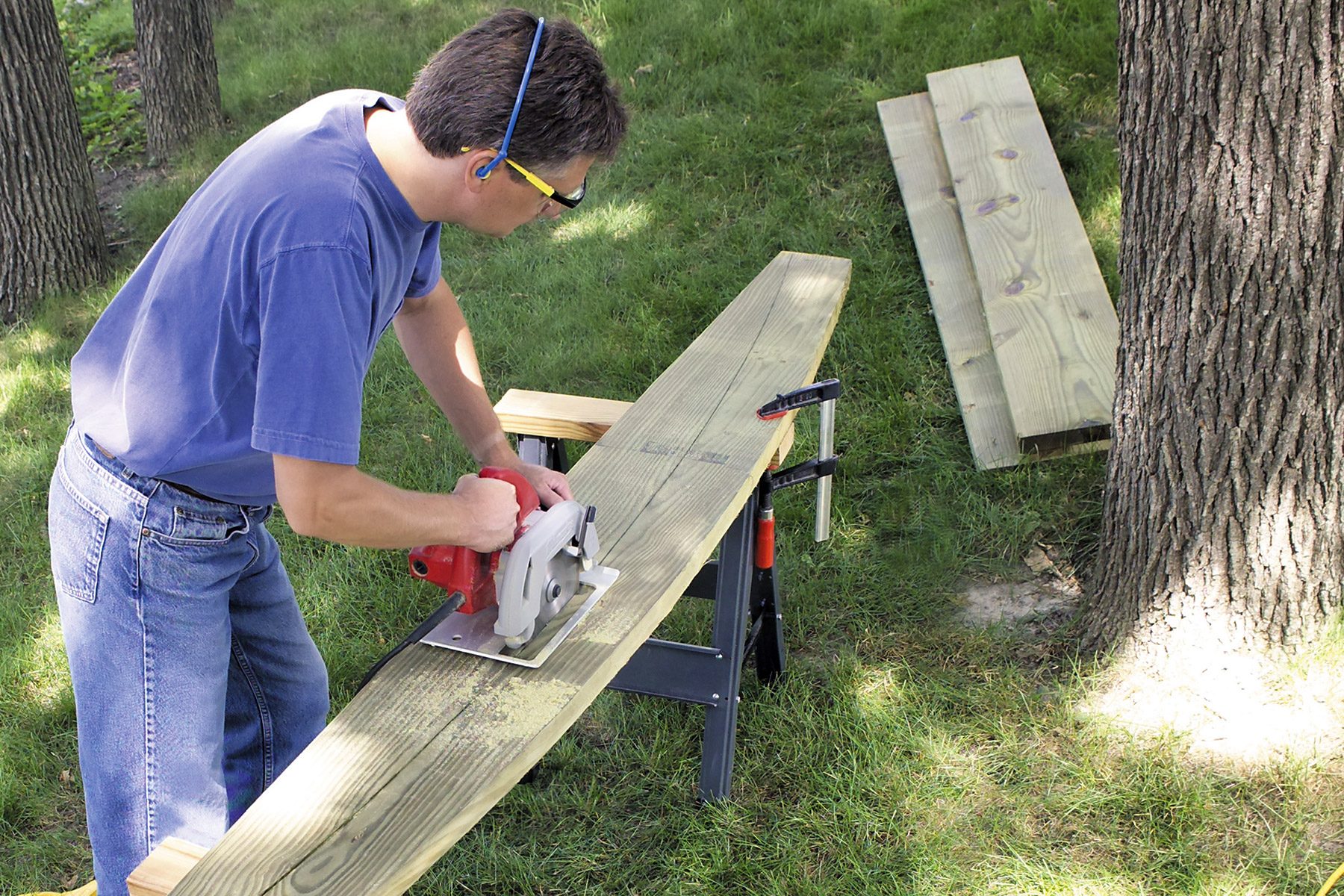 A person wearing a blue shirt, sunglasses, and ear protection uses a circular saw to cut a large wooden plank on a portable workbench outdoors. Two additional planks are on the grass nearby, and the setting appears to be a grassy backyard.