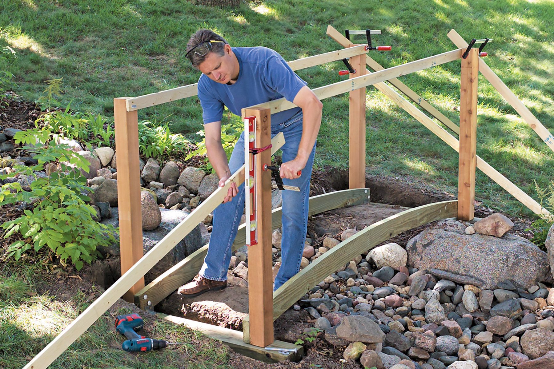 A man wearing a blue shirt and jeans is building a wooden bridge in a garden area. He is using a hand tool to secure a vertical post of the bridge frame. The surrounding area includes rocks and greenery. Various clamps and tools are visible in the work area.