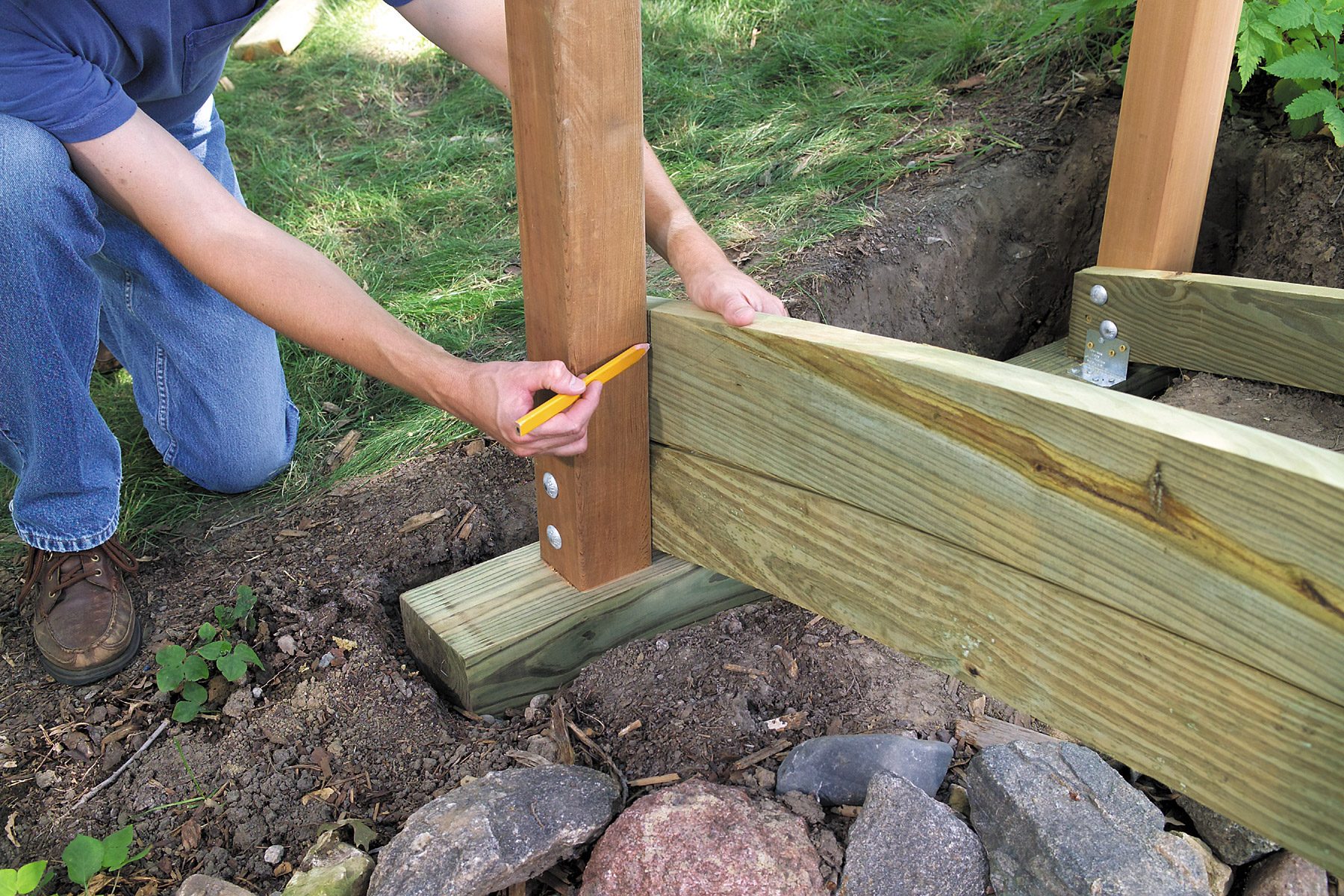 A person is using a carpenter's pencil to mark a wooden post. The post is part of a larger wooden structure with metal brackets, set in a grassy area with rocks and dirt at the base. The scene appears to be part of a construction or woodworking project.