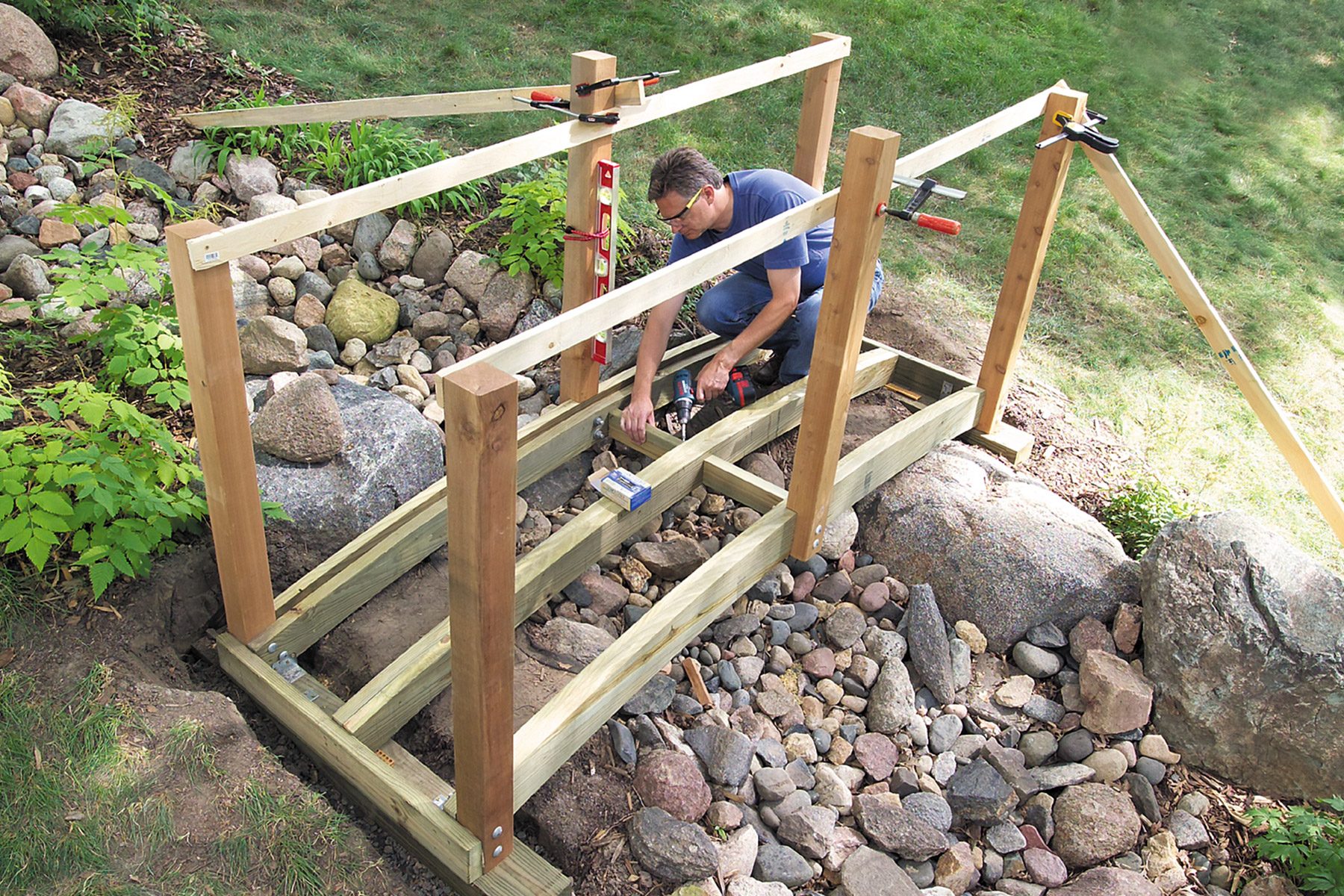 A person in a blue shirt is constructing a wooden footbridge over a rocky area. The bridge frame is partially complete with vertical posts and horizontal beams. Various tools and clamps are visible, with a grassy area in the background.