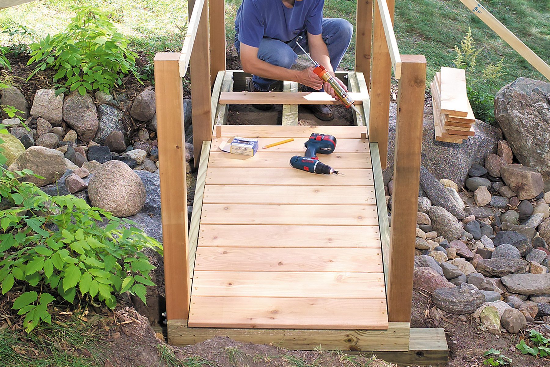 A person wearing a blue shirt is constructing a wooden bridge, kneeling on unfinished decking. Various tools, including a drill, clamps, and a pencil, are placed around them. In the background, there are rocks, greenery, and a partially built handrail.