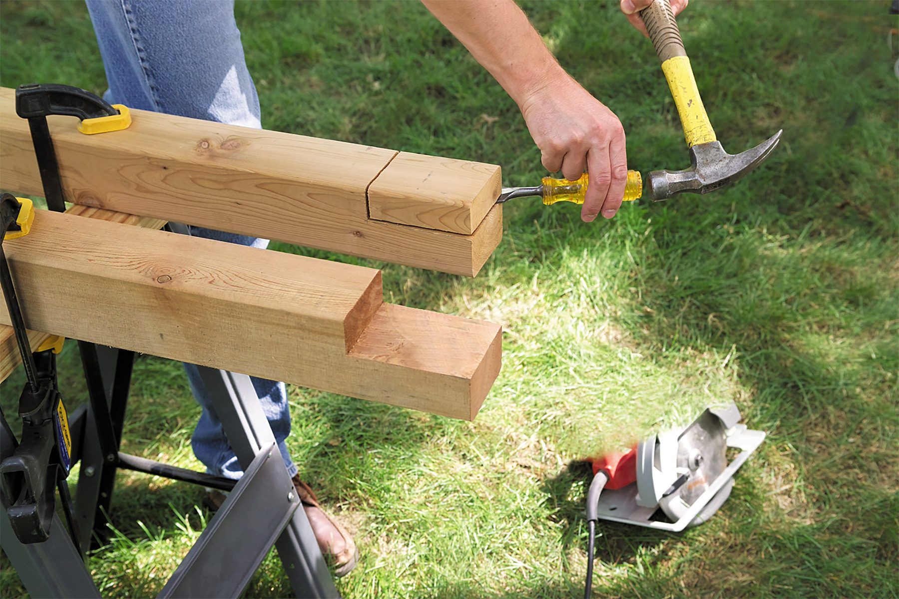 A person uses a chisel and hammer to cut precise notches in wooden beams outdoors. The beams rest on a saw horse, and a circular saw is visible on the ground nearby. The grass and legs of the individual can be seen in the background.