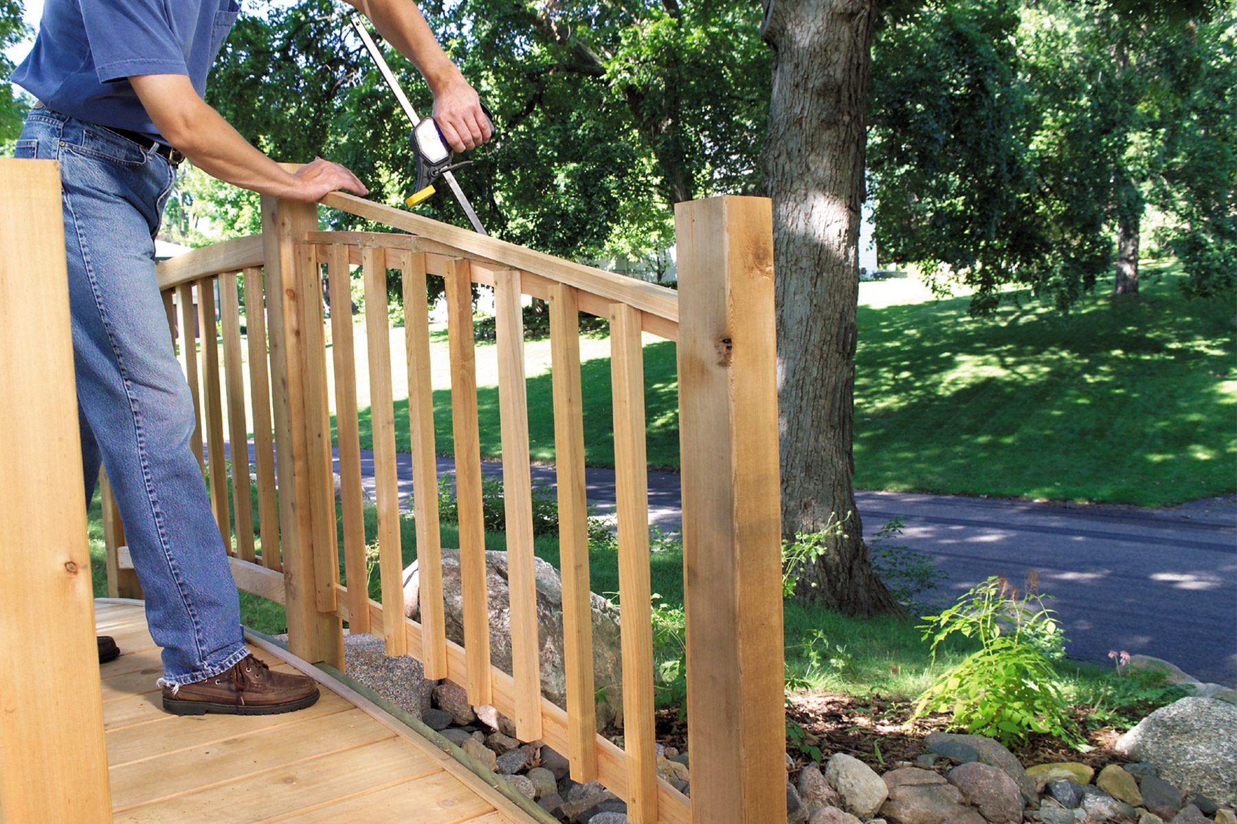 A person in jeans is using a power drill to assemble or repair a wooden railing on a deck. The background features a lush, green outdoor setting with trees and a pathway. Rocks are visible underneath the deck.