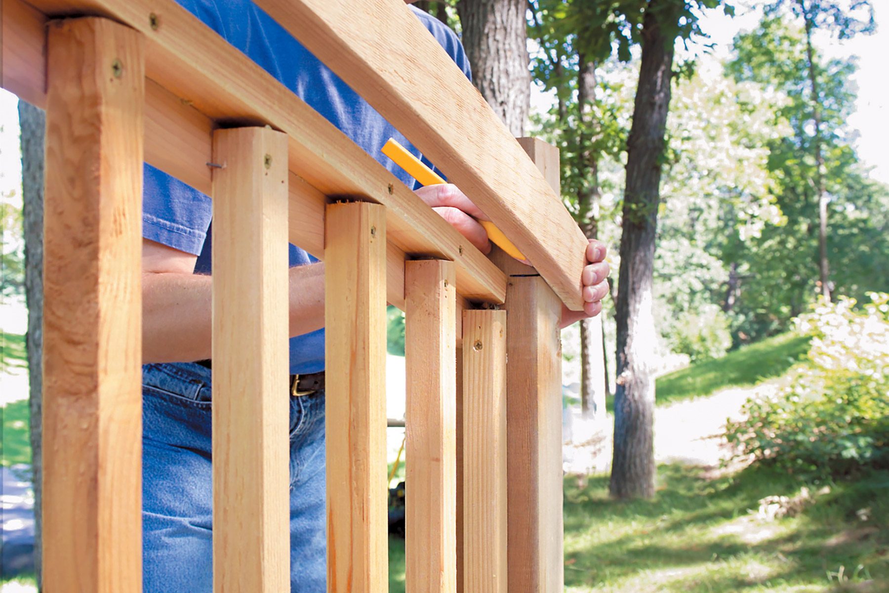 Person in blue shirt holding a wooden top rail in place while using a yellow measuring tool to mark it, constructing or repairing a wooden railing outdoors with trees in the background.