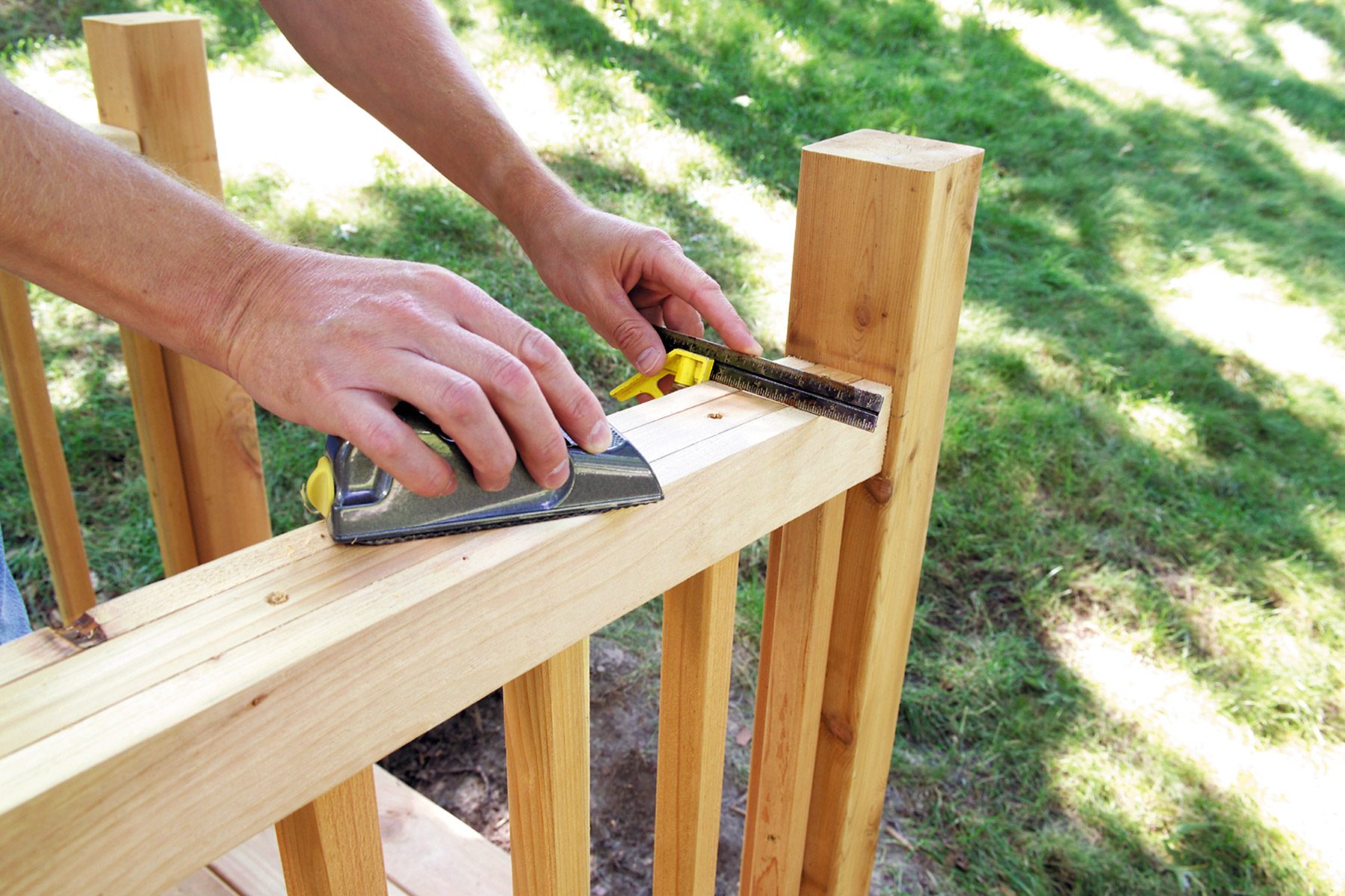 A person measures a wooden railing with a tape measure and holds a square tool against it. The railing is part of an outdoor structure, and the background shows a grassy area bathed in sunlight. The wood appears newly cut and unfinished.