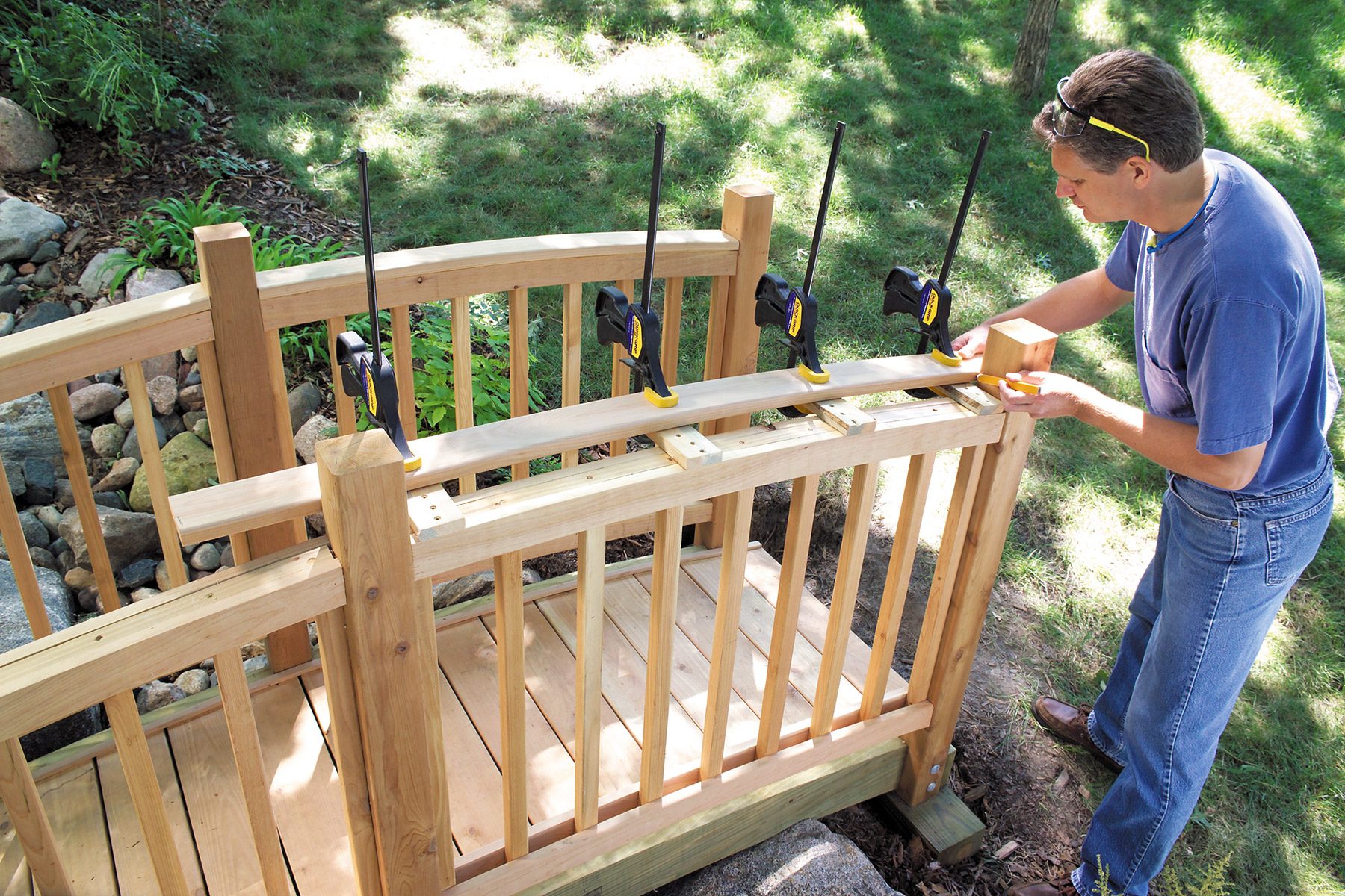 A person works on building a wooden railing for an outdoor staircase. Clamps are securing the handrail in place. The structure is surrounded by green grass and trees, indicating a garden or backyard setting. The railing appears to be nearly completed.