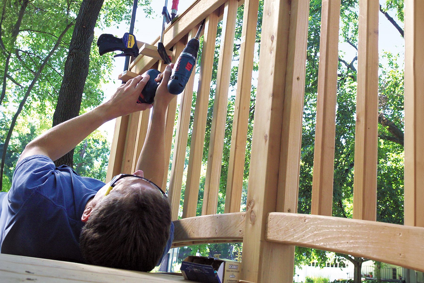 A person lying on their back wearing safety goggles is using a power drill to secure a clamp on a wooden structure. The scene appears to be set outdoors in a sunny area with trees in the background. There are tools and materials around the person.