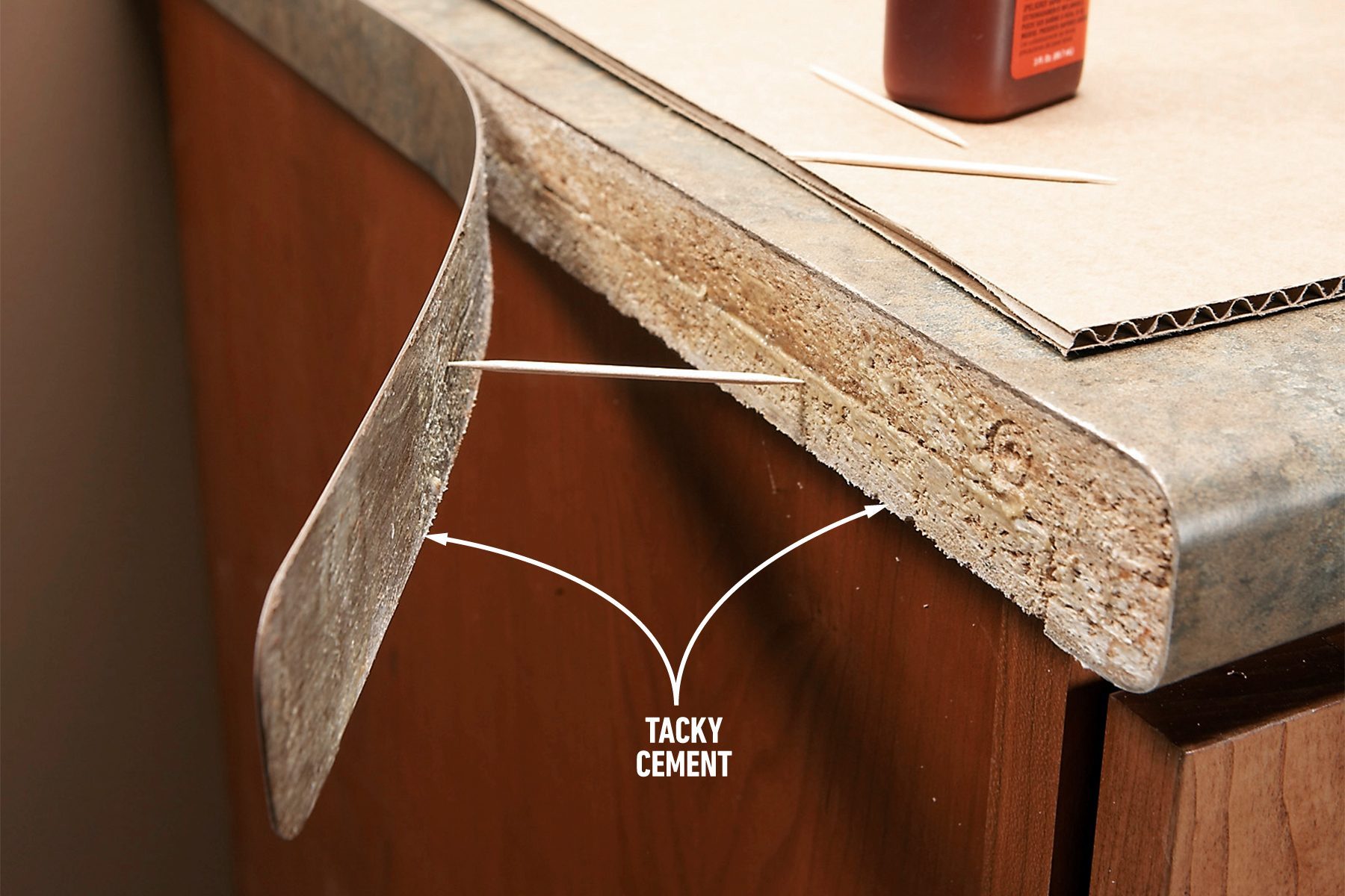 A piece of laminate countertop is lifted at the corner to show a layer of tacky cement beneath it. The edge of the laminate curves upward, while the wooden base and a bottle of cement sit on top of the counter. Two arrows point to the tacky cement layer below.