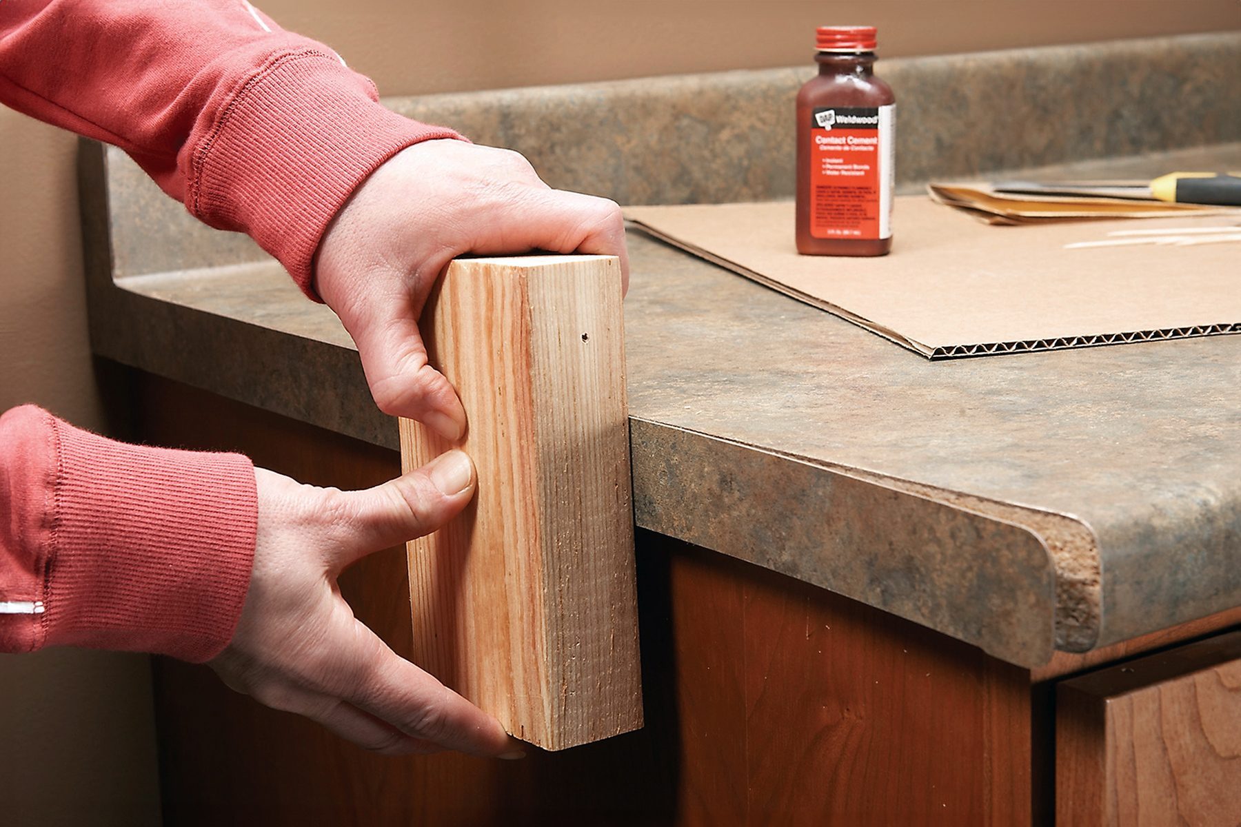 A person in a red sweatshirt is holding a piece of wood horizontally in front of a countertop with a wooden edge. A bottle of carpentry adhesive is visible on the countertop along with a piece of cardboard and a putty knife.