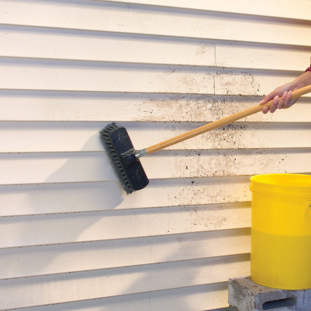 A person scrubs the dirty siding of a house with a long-handled brush. There is a yellow bucket on a cinder block nearby, presumably holding water or cleaning solution to assist with the cleaning. The house siding has horizontal lines.