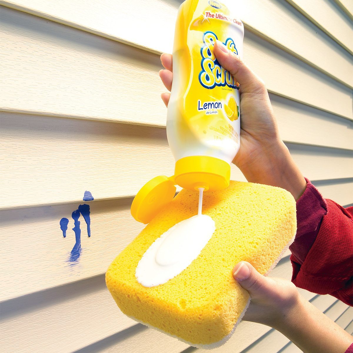 A person is squeezing a bottle of liquid cleaner labeled "Soft Scrub Lemon" onto a yellow sponge. The person is using the sponge to clean a blue stain off beige vinyl siding. The person's other hand holds the sponge against the siding.