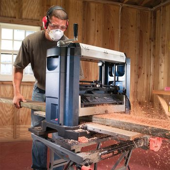A person wearing safety goggles, ear protection, and a dust mask is operating a planer in a wooden workshop. They are feeding a long piece of wood through the machine, with wood shavings being produced and scattered around.