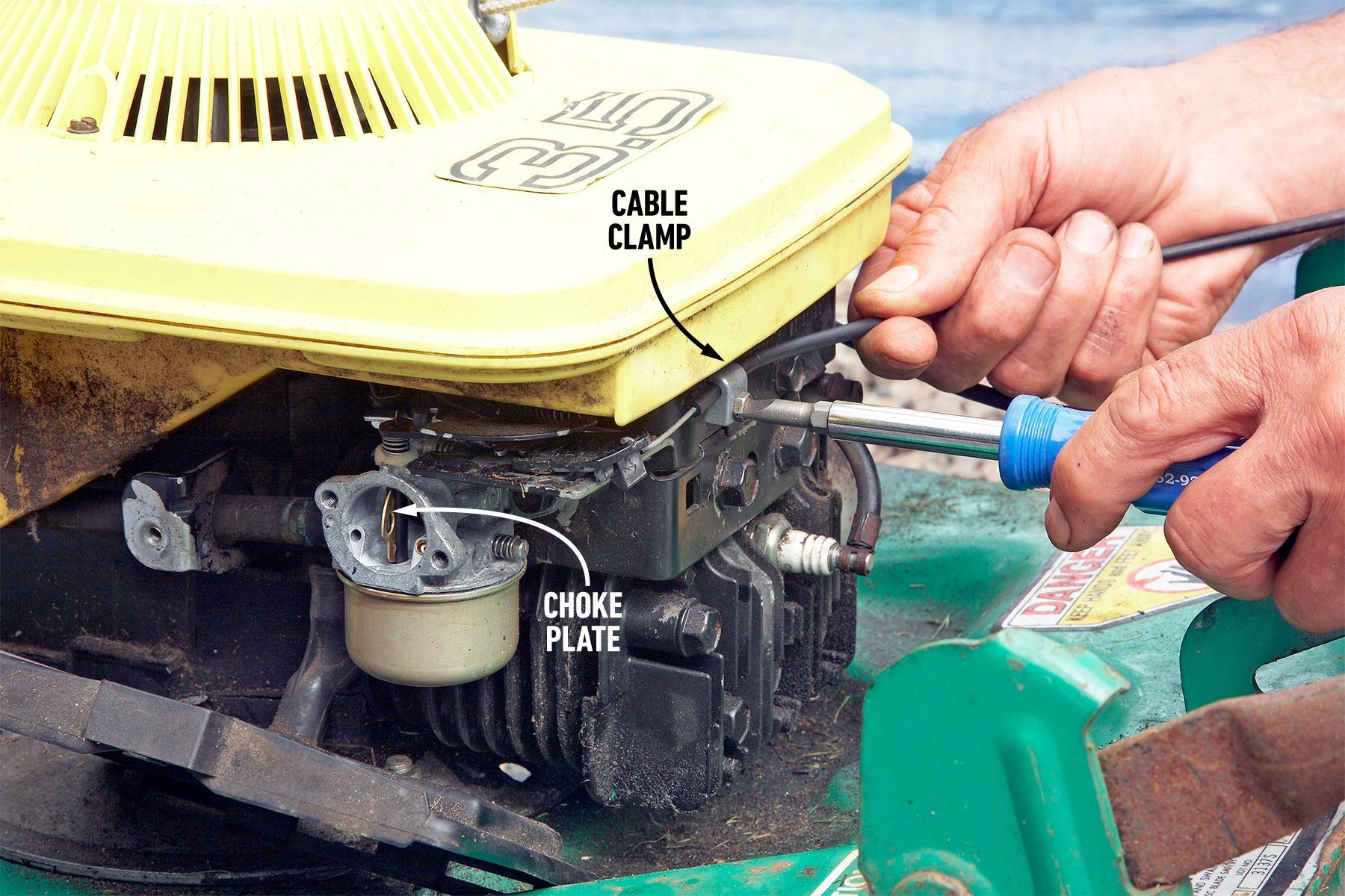 Close-up of a person's hand using a screwdriver to adjust a cable clamp near the choke plate of a small engine. The engine has a yellow cover, and the green base is visible in the foreground. Labels identify the cable clamp and the choke plate.