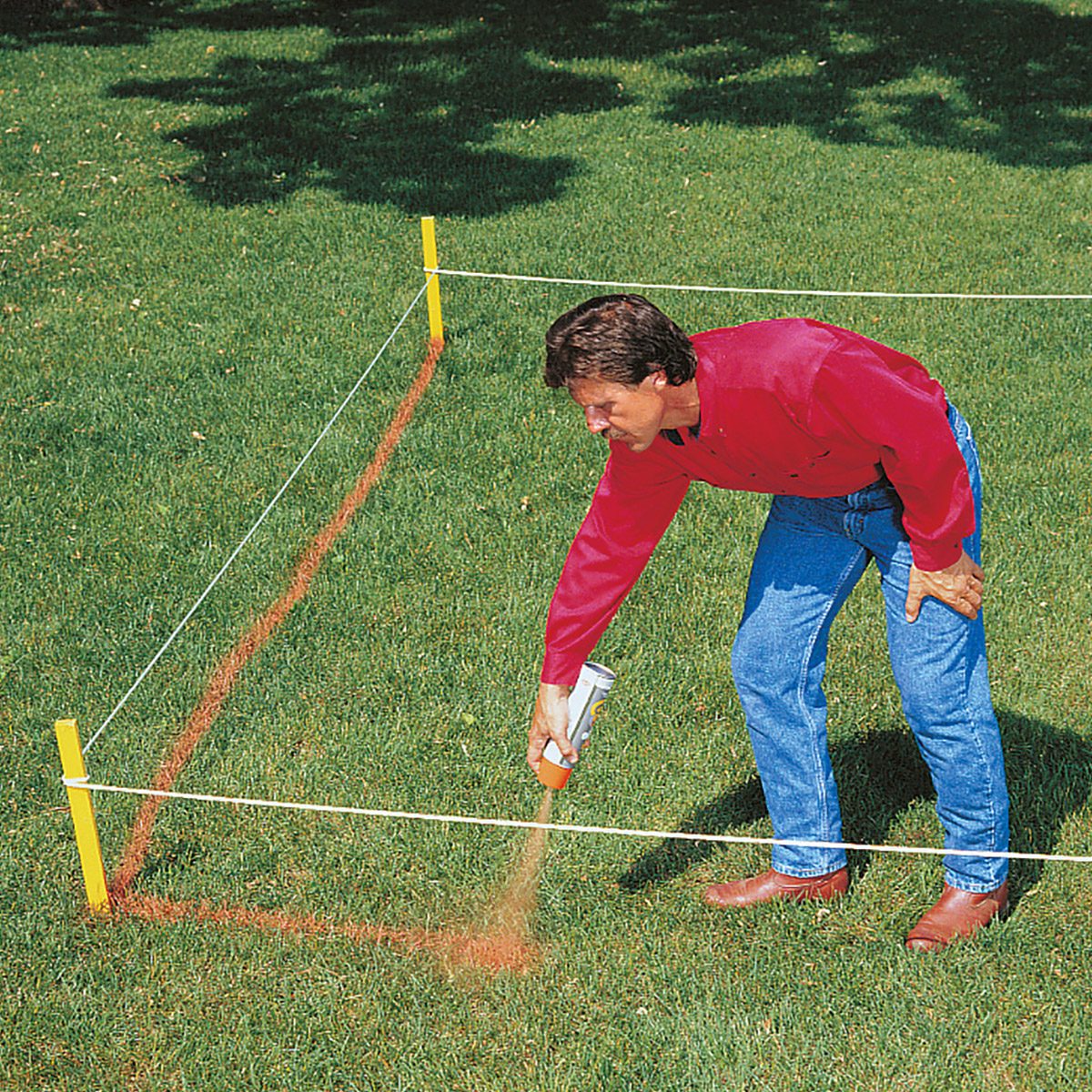 A person wearing a red long-sleeve shirt, blue jeans, and brown shoes is bent over, spray-painting lines on a grassy area marked by string tied to four yellow stakes. The person appears to be creating a rectangular outline on the grass.