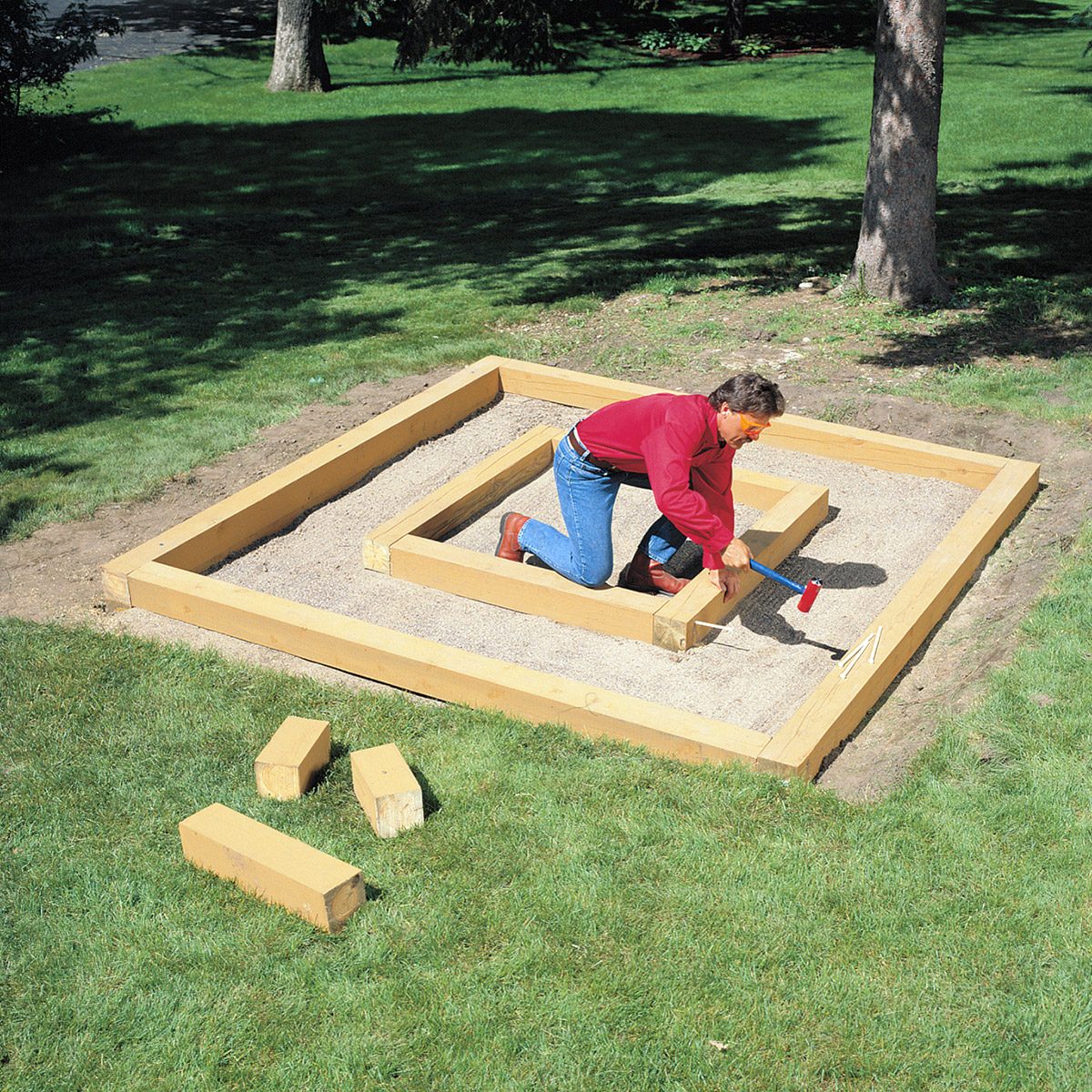 A person is assembling a wooden structure on a patch of dirt within a grassy area. They are using a hammer to secure the wood pieces together. There are extra wooden blocks on the grass nearby, and trees can be seen in the background.