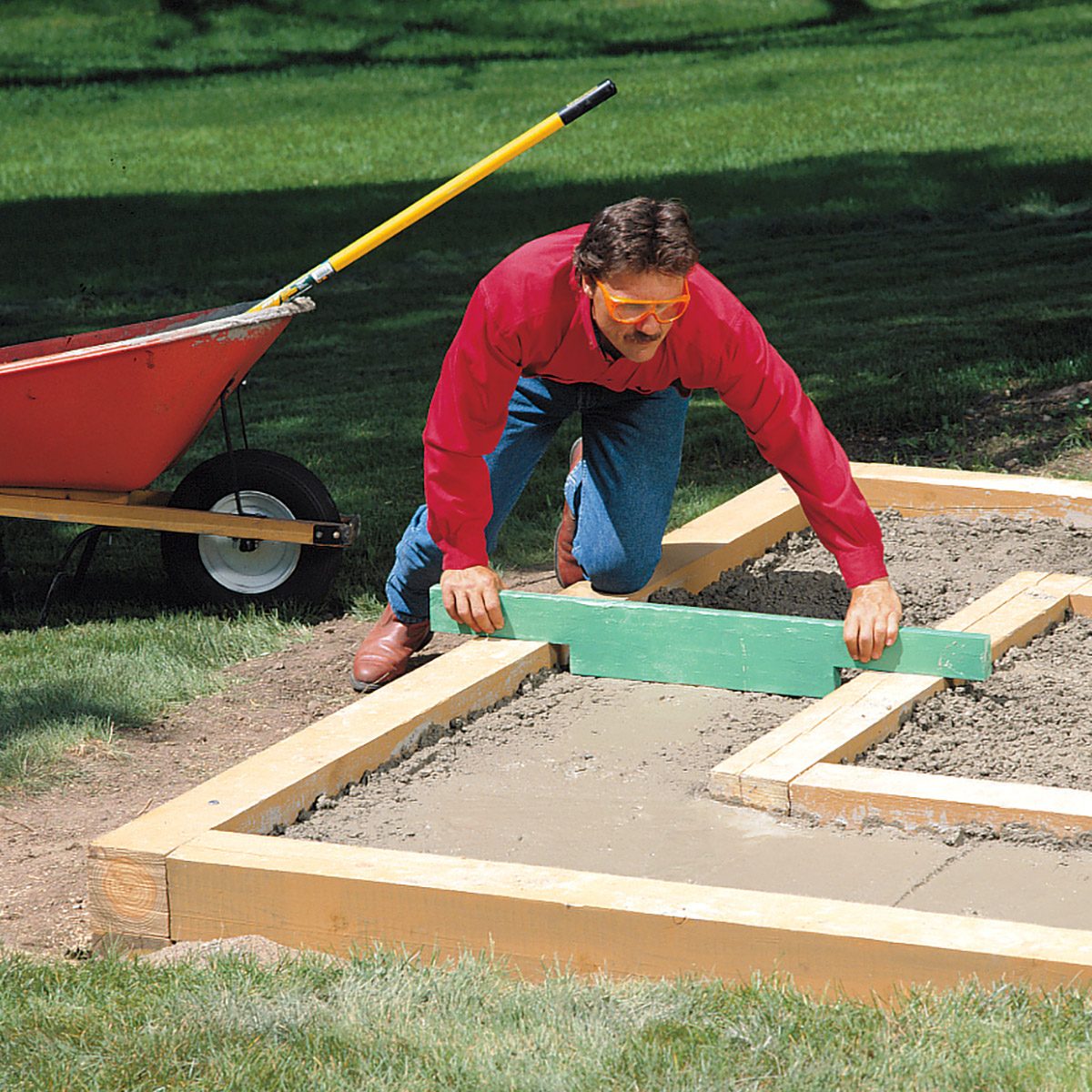 A person wearing a red shirt and blue jeans is leveling concrete within a wooden frame using a green tool. A red wheelbarrow is nearby on a grassy area. The background features a green lawn and partial shade.