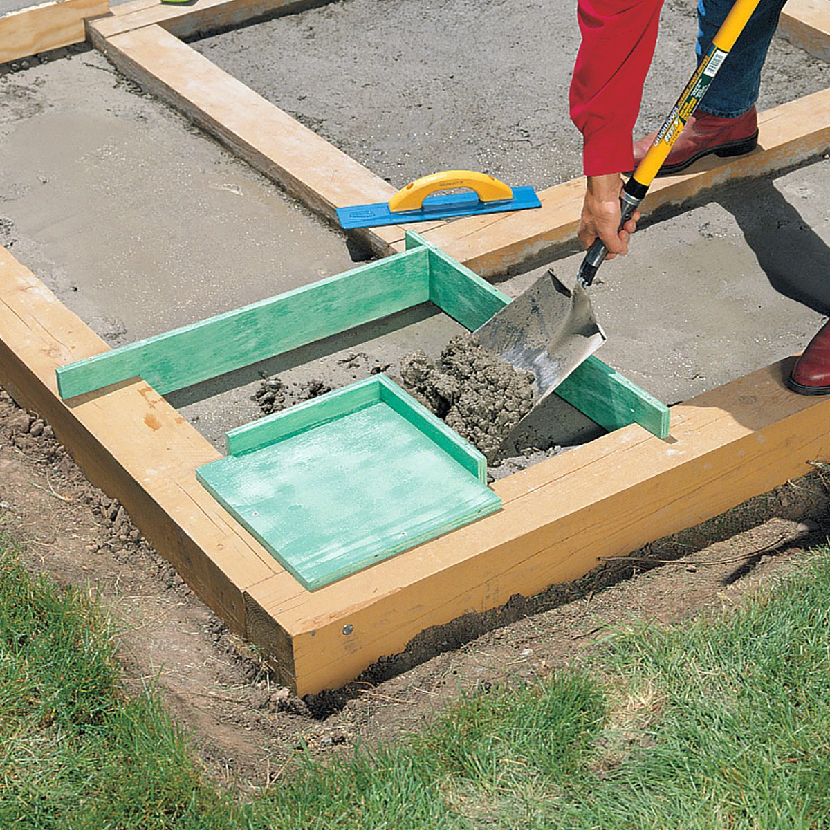 A person is using a tool to spread concrete inside a wooden frame. There's a green square mold within the frame, and a trowel in the worker's hand. The frame is placed on grass, partially filled with gravel or concrete mix.