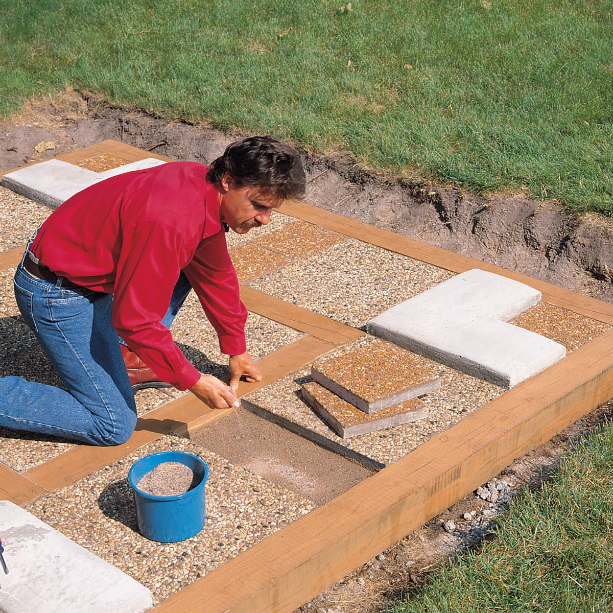 A man in a red shirt and blue jeans kneels on the ground while fitting pavers and pea gravel into a wooden grid framework. A blue bucket with sand is next to him, and the work is taking place on a grassy lawn.