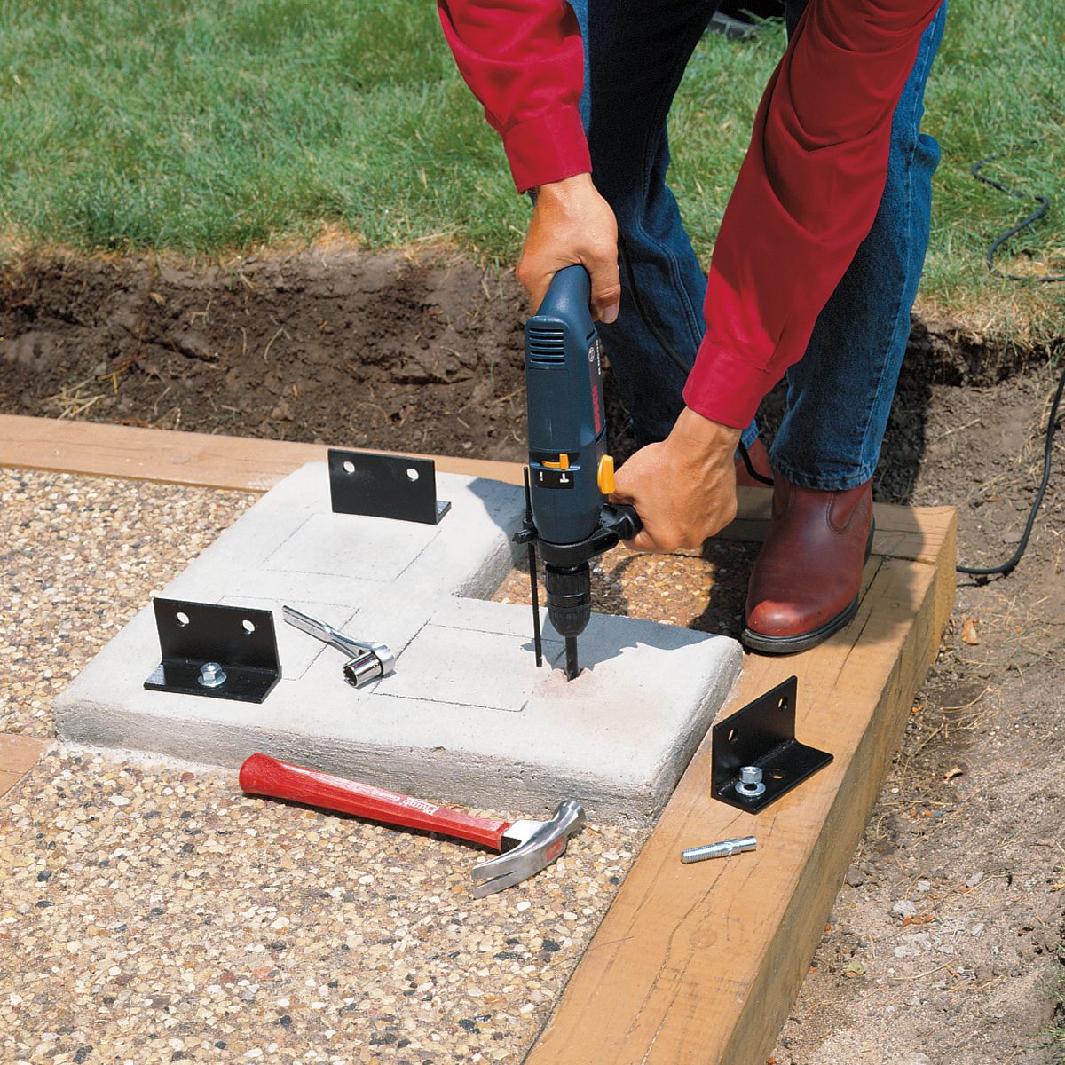 A person in a red shirt and blue jeans is using a power drill on a concrete base with metal brackets. Nearby on the ground are a hammer, screws, and a wrench. The scene is set outdoors on a lawn area.