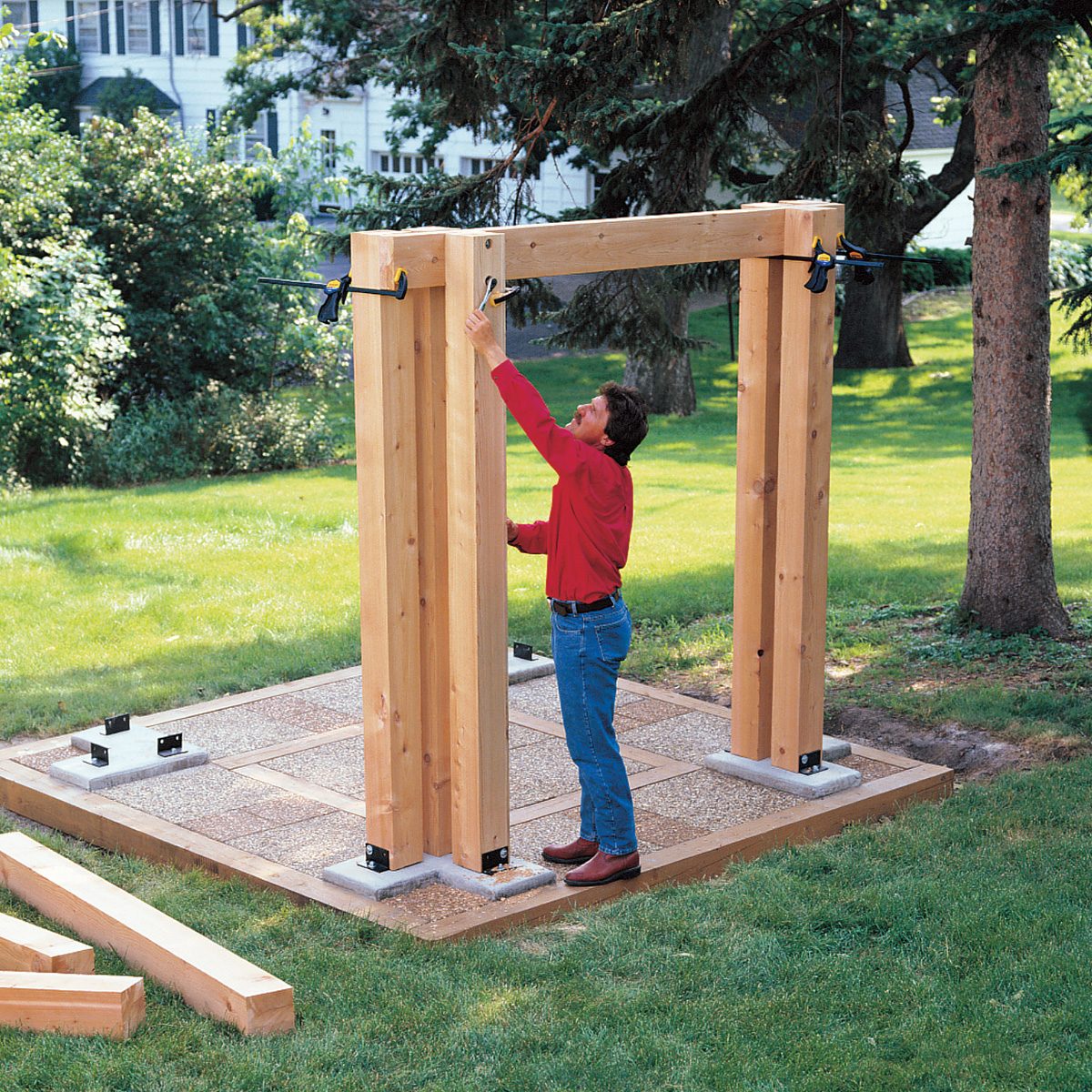 A person in a red shirt and blue jeans is installing a wooden pergola frame in a grassy backyard. The structure is partially assembled on a tiled base, surrounded by trees and greenery. Some wooden beams are lying on the ground nearby.