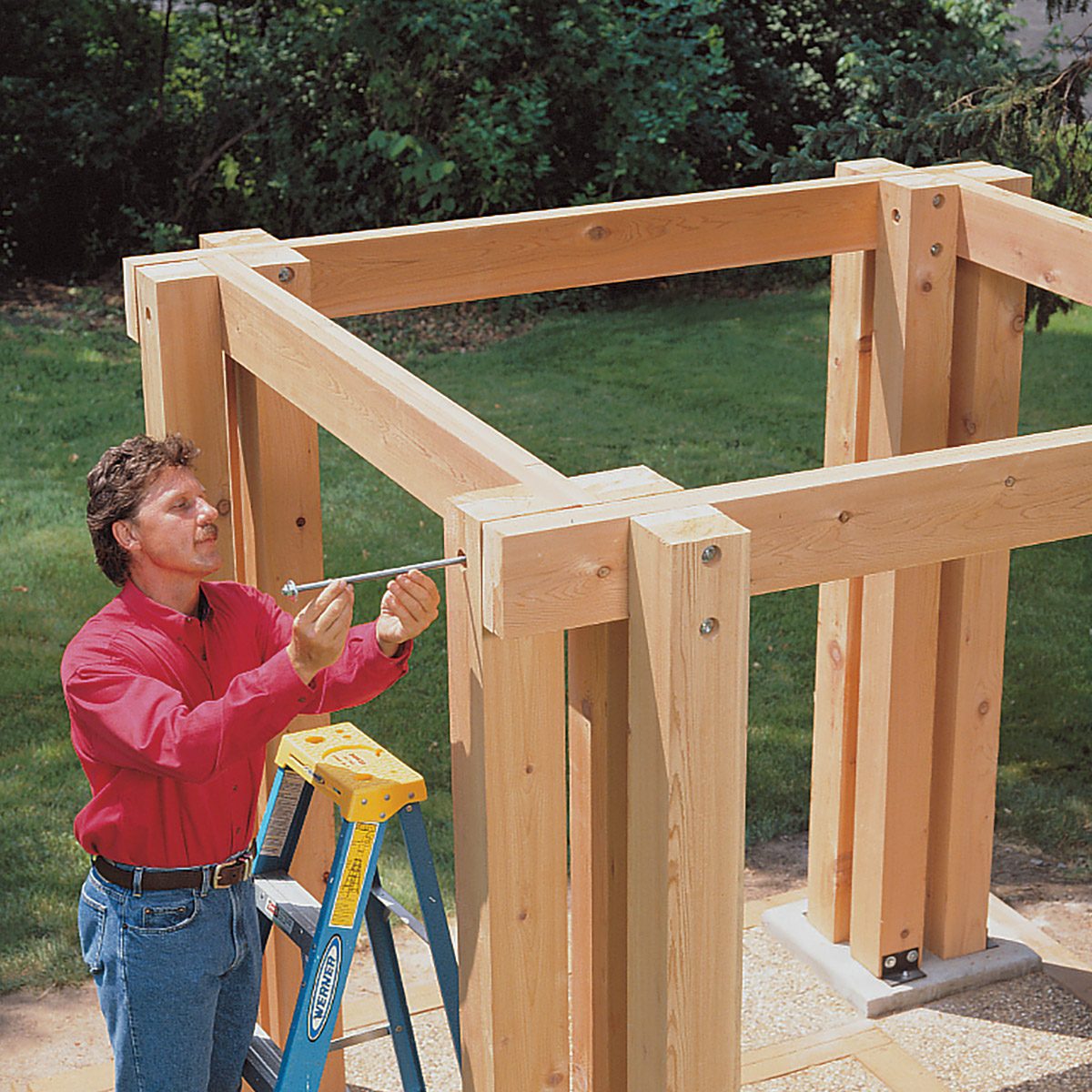 A person in a red shirt is standing on a blue ladder using a level tool to check the alignment of a partially constructed wooden pergola in a garden. The structure consists of large wooden beams secured together with bolts, and there is greenery in the background.