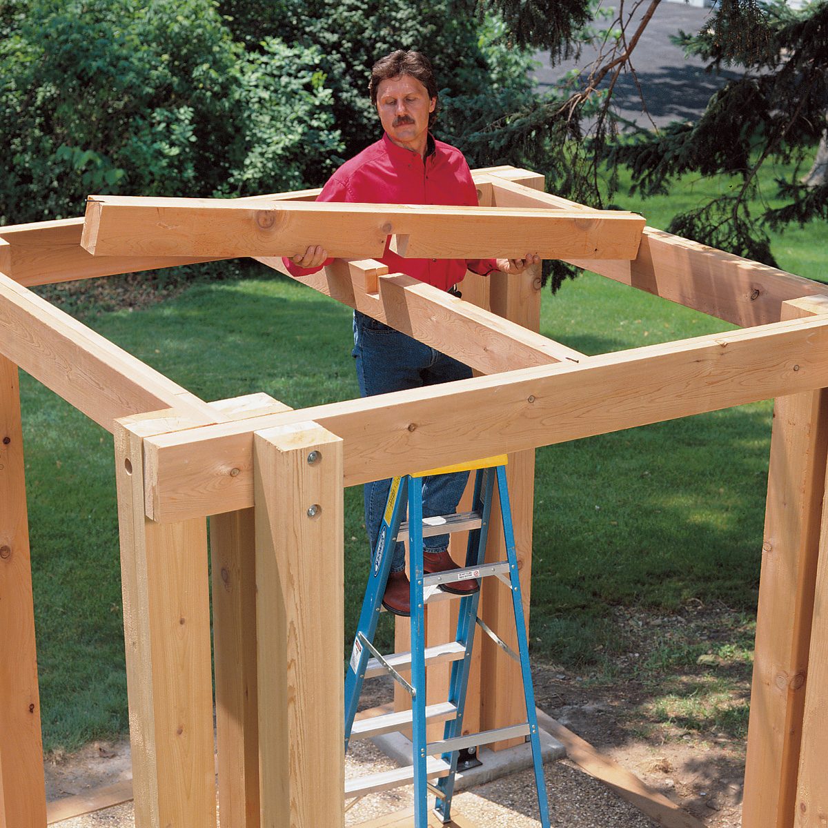 A person in a red shirt stands on a ladder, constructing a wooden outdoor structure in a grassy area with trees. The incomplete structure consists of large, light-colored beams and posts, forming a frame.