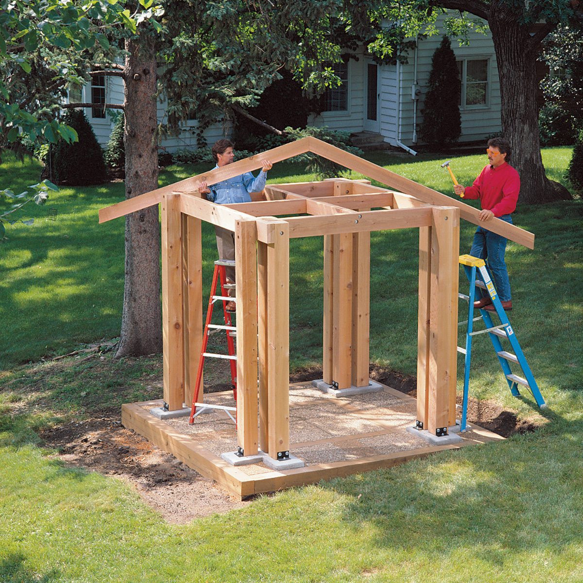 Two people are constructing a wooden shed frame outdoors. The frame consists of vertical and horizontal beams forming the structure's skeleton. One person on a red ladder and another on a blue ladder work on attaching pieces. 