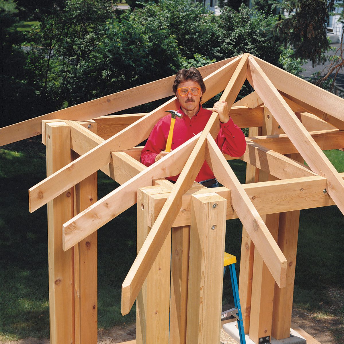 A person in a red shirt stands within a partially constructed wooden structure outdoors. They are holding a yellow hammer and appear to be focused on the construction task. The framework suggests the structure will be a small building or shed. Trees and grass are visible in the background.